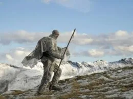 Ein Jäger in winterlicher Tarnkleidung bei der Jagd in den verschneiten Bergen, mit einem langen Stab in der Hand, umgeben von der majestätischen Winterlandschaft.