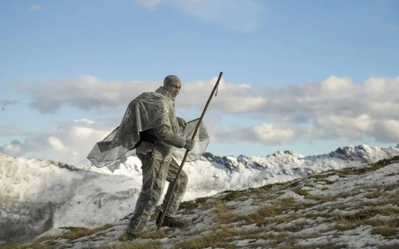 Ein Jäger in winterlicher Tarnkleidung bei der Jagd in den verschneiten Bergen, mit einem langen Stab in der Hand, umgeben von der majestätischen Winterlandschaft.