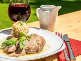 Wildschweinschlögel mit Kräuterknödel, serviert mit Sauce und Rotwein auf einem Holztisch © Martin Grasberger