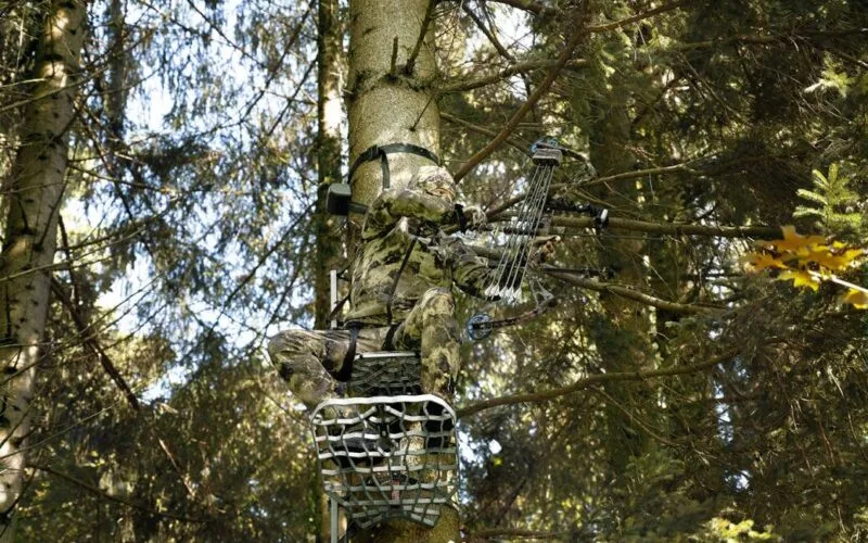 Weidgerechte Bogenjagd - Ein Jäger im Tarnanzug sitzt auf einem Hochsitz, bereit für die Bogenjagd im Wald.