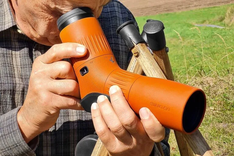 Jäger oder Naturbeobachter nutzt ein hochwertiges Spektiv mit brauner Gummiarmierung, abgestützt auf einem Jagdstock im freien Gelände