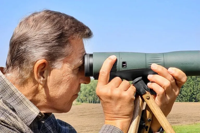 Mann beobachtet die Landschaft mit einem hochwertigen Spektiv, abgestützt auf einem Jagdstock für eine ruhige Handhabung
