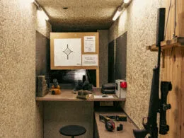 Ein Jagdschießstand mit einer aufgesetzten Zielscheibe, einer Jagdbüchse mit Schalldämpfer an der Wandhalterung, Munition und Schießausrüstung auf dem Tisch.