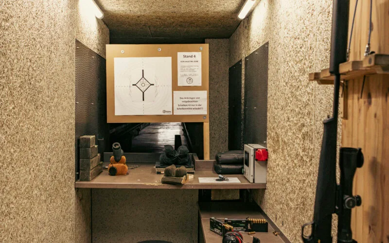Ein Jagdschießstand mit einer aufgesetzten Zielscheibe, einer Jagdbüchse mit Schalldämpfer an der Wandhalterung, Munition und Schießausrüstung auf dem Tisch.