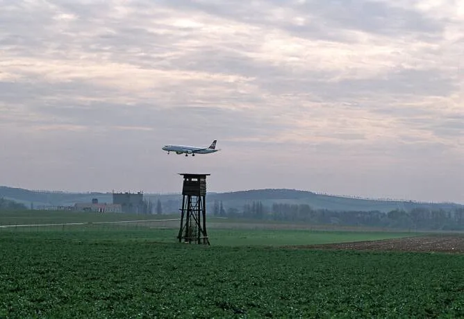 Ein Flugzeug fliegt in niedriger Höhe über ein landwirtschaftliches Feld mit einem Hochsitz im Vordergrund.