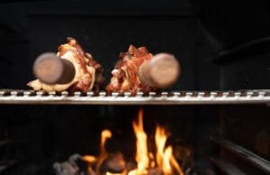 Nahaufnahme von zwei Wildfleischspießen auf einem Grillrost über offenem Feuer, im Hintergrund lodernde Flammen.