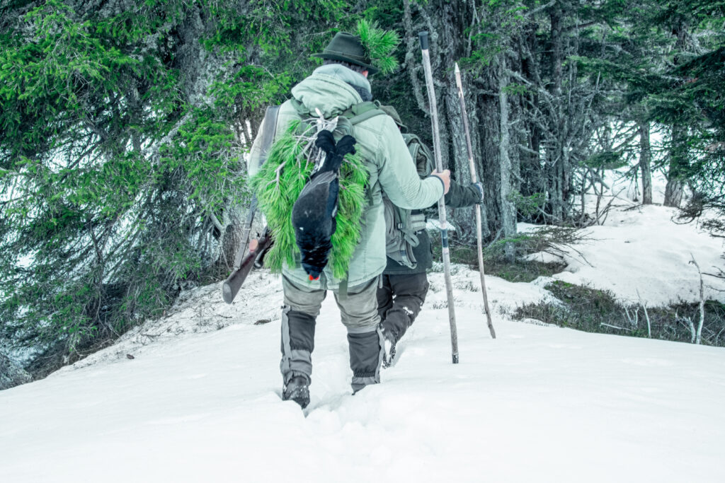 Zwei Jäger beim Aufstieg im Schnee, einer trägt einen erlegten Birkhahn im Rucksack
