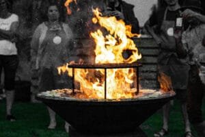 Offenes Feuer in einer Feuerschale mit Grillaufsatz bei einem Grillkurs im Freien, umringt von Menschen.