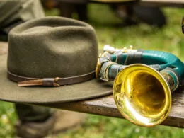 Ein traditionelles Jagdhorn mit grün umwickeltem Korpus und goldener Schallöffnung liegt auf einer Holzbank neben einem grünen Jagdhut mit braunem Lederband. Die Szene vermittelt eine rustikale, jagdliche Atmosphäre im Freien.