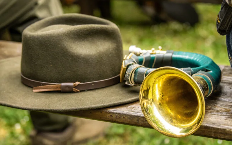 Ein traditionelles Jagdhorn mit grün umwickeltem Korpus und goldener Schallöffnung liegt auf einer Holzbank neben einem grünen Jagdhut mit braunem Lederband. Die Szene vermittelt eine rustikale, jagdliche Atmosphäre im Freien.
