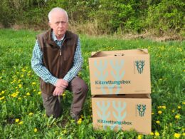 Älterer Jäger kniet auf einer Wiese neben zwei Kitzrettungsboxen des NÖ Jagdverbands mit dem Aufdruck „Dem Wild verpflichtet“.