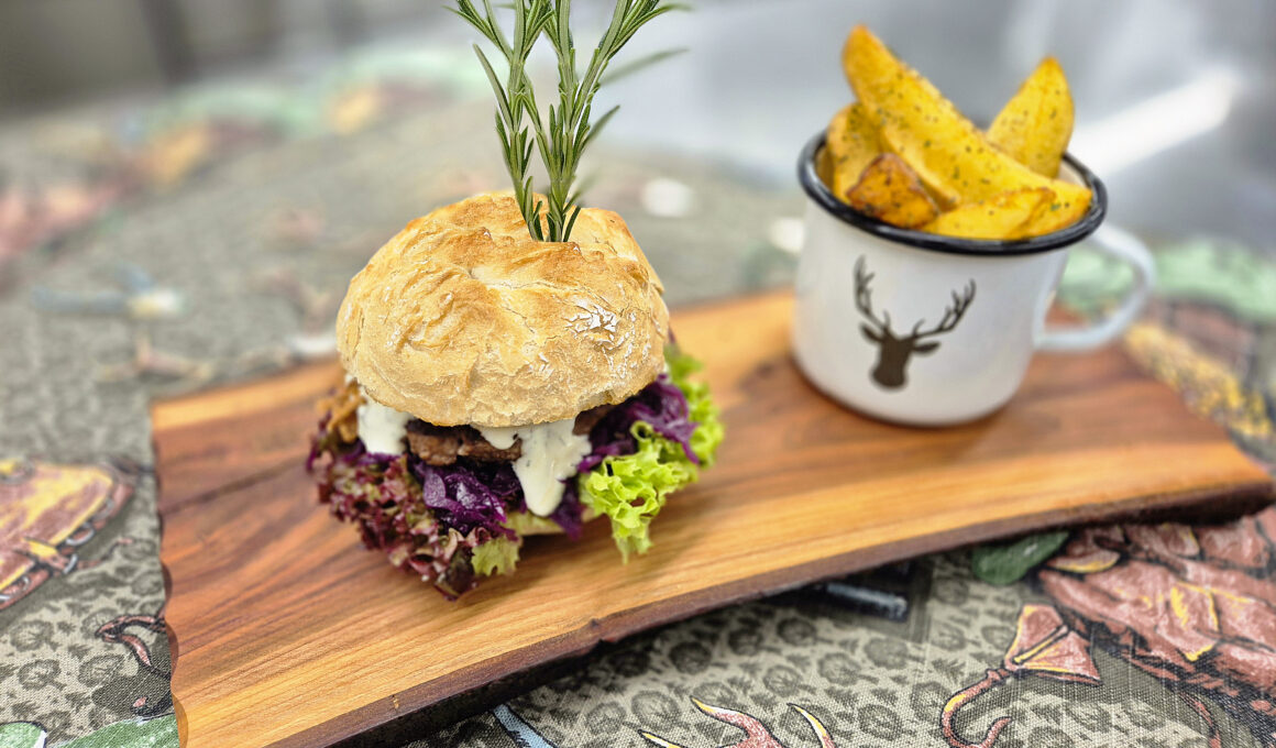 Saftiger Wild-Burger mit Rotkraut, Schnittlauchsauce und Rosmarin-Garnitur, serviert mit knusprigen Potato Wedges im Emaille-Becher
