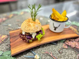 Saftiger Wild-Burger mit Rotkraut, Schnittlauchsauce und Rosmarin-Garnitur, serviert mit knusprigen Potato Wedges im Emaille-Becher