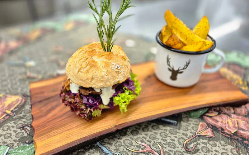 Saftiger Wild-Burger mit Rotkraut, Schnittlauchsauce und Rosmarin-Garnitur, serviert mit knusprigen Potato Wedges im Emaille-Becher