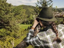 Frau mit grünem Jagdhut beobachtet mit Fernglas die Natur von einem Hochstand aus