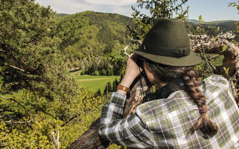 Frau mit grünem Jagdhut beobachtet mit Fernglas die Natur von einem Hochstand aus
