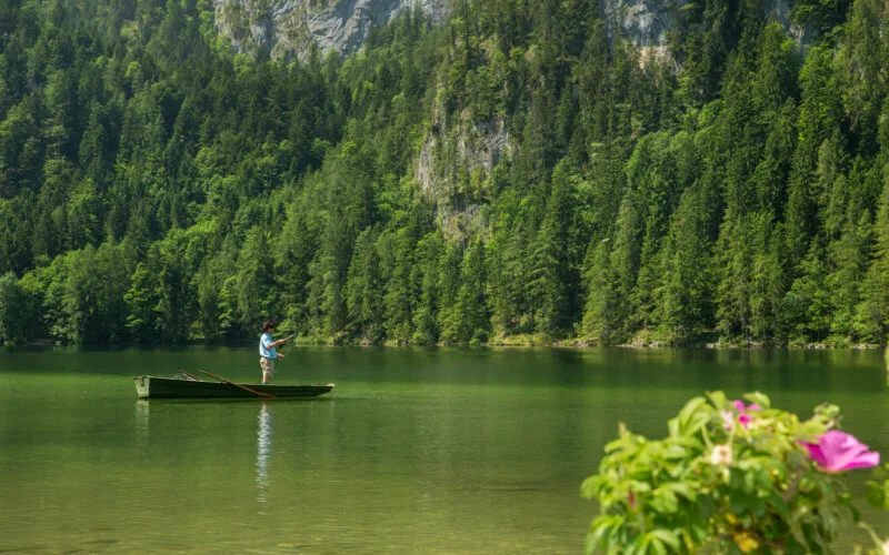 Fliegenfischer im Boot auf klarem Bergsee vor bewaldetem Felsmassiv