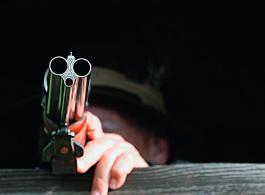 Lauf eines Drillings im Vordergrund – Jäger im Ansitz zielt konzentriert aus der Deckung
