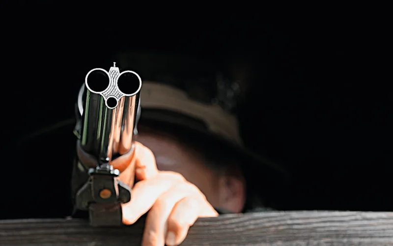 Lauf eines Drillings im Vordergrund – Jäger im Ansitz zielt konzentriert aus der Deckung
