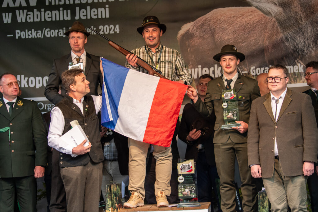 Der französische Sieger der Hirschruf-Europameisterschaft 2024 steht mit Gewehr und Nationalflagge auf dem Podest, flankiert von Mitbewerbern in Jagduniformen.