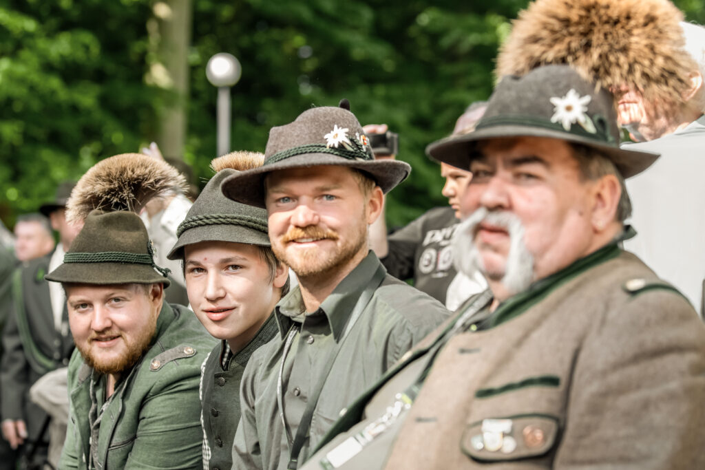 Vier Jäger in traditioneller Tracht mit Gamsbart und Edelweiß-Anstecker sitzen lächelnd in einer Zuschauerreihe bei der Hirschruf-EM 2024 in Polen.